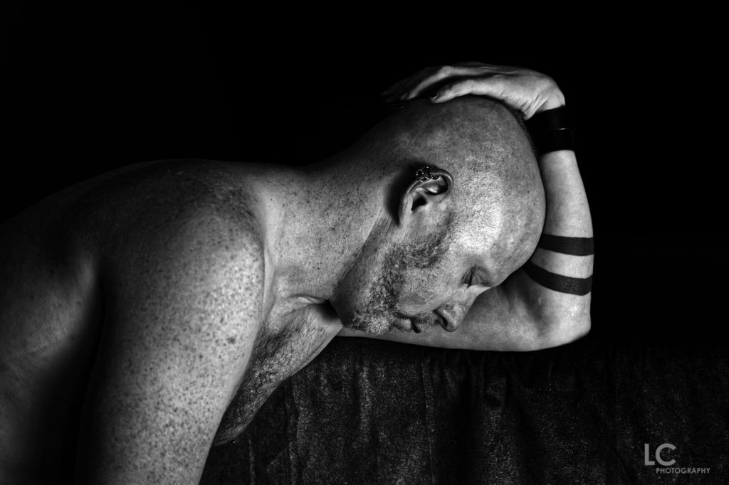 Shirtless male leaning forward on bench, face downwardss, holding head on bicep  of bent arm.