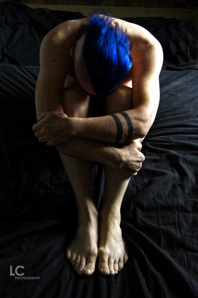 implied nude of male model seated with head bowed and arms wrapped around legs